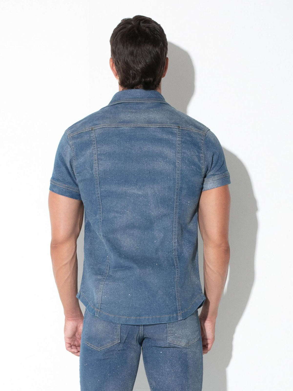 A man viewed from behind wearing a blue denim short-sleeve button-up shirt and matching blue jeans against a white background.