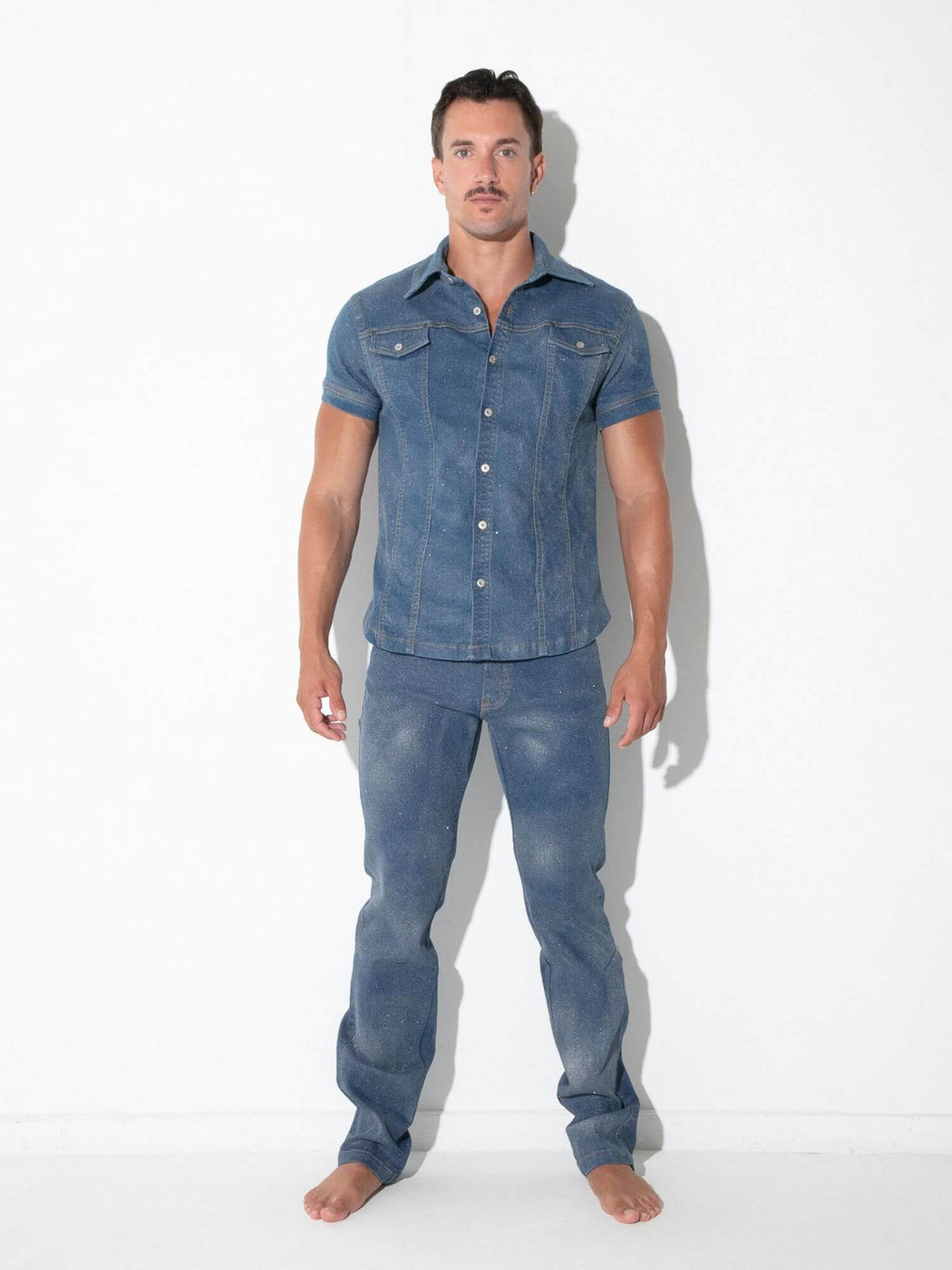 A man modeling a matching blue denim short-sleeve button-up shirt and jeans set against a plain white studio background.