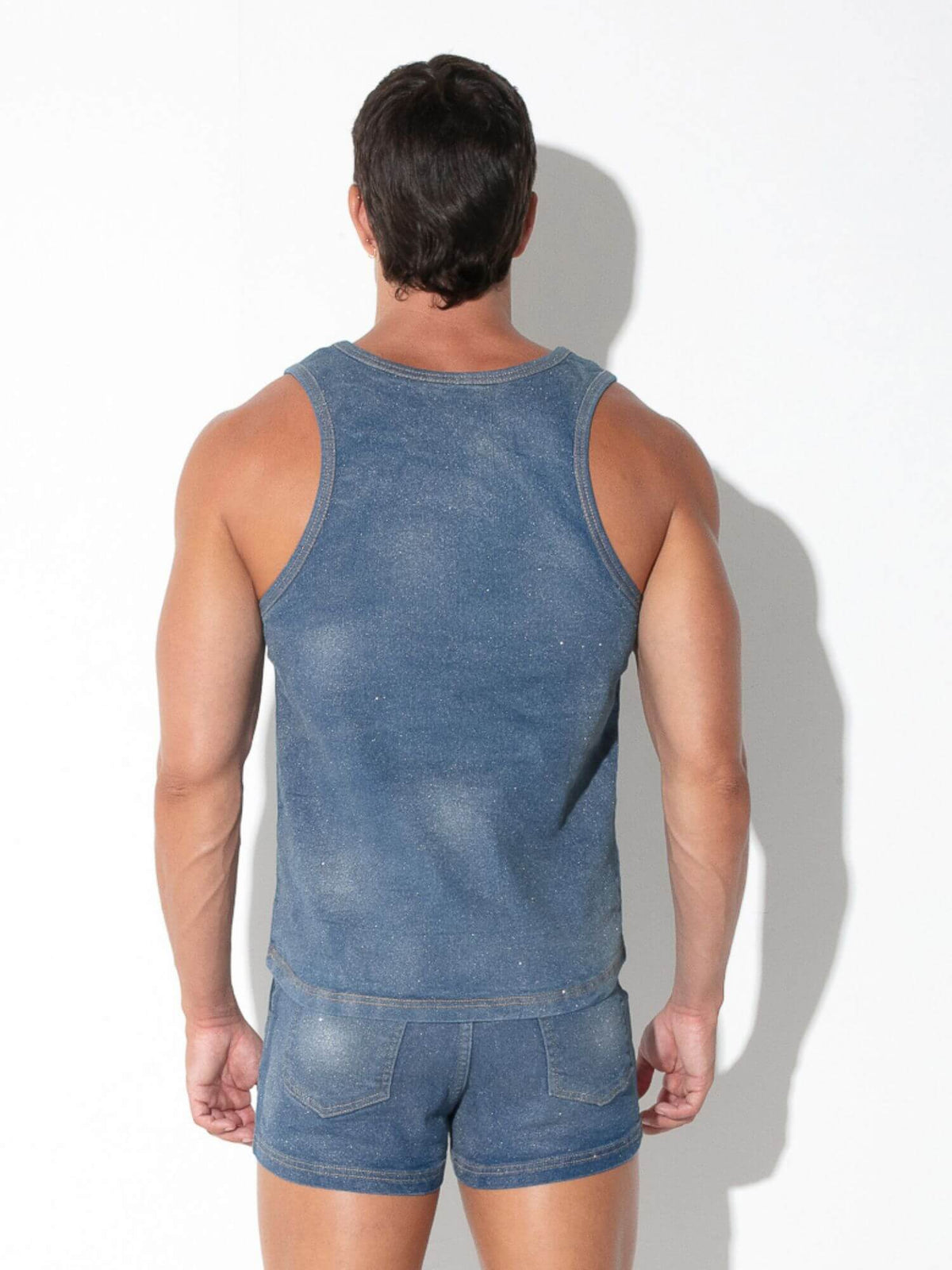A man viewed from behind wearing a faded blue denim tank top and matching denim shorts against a white wall with a soft shadow.