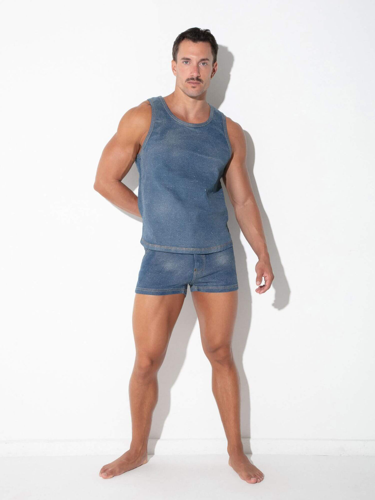 A muscular man modeling a matching blue denim tank top and shorts set against a plain white studio background.