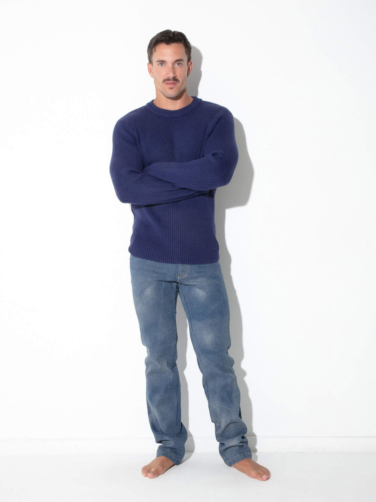 A man standing barefoot against a white studio wall, wearing a dark navy ribbed knit sweater and light blue jeans.