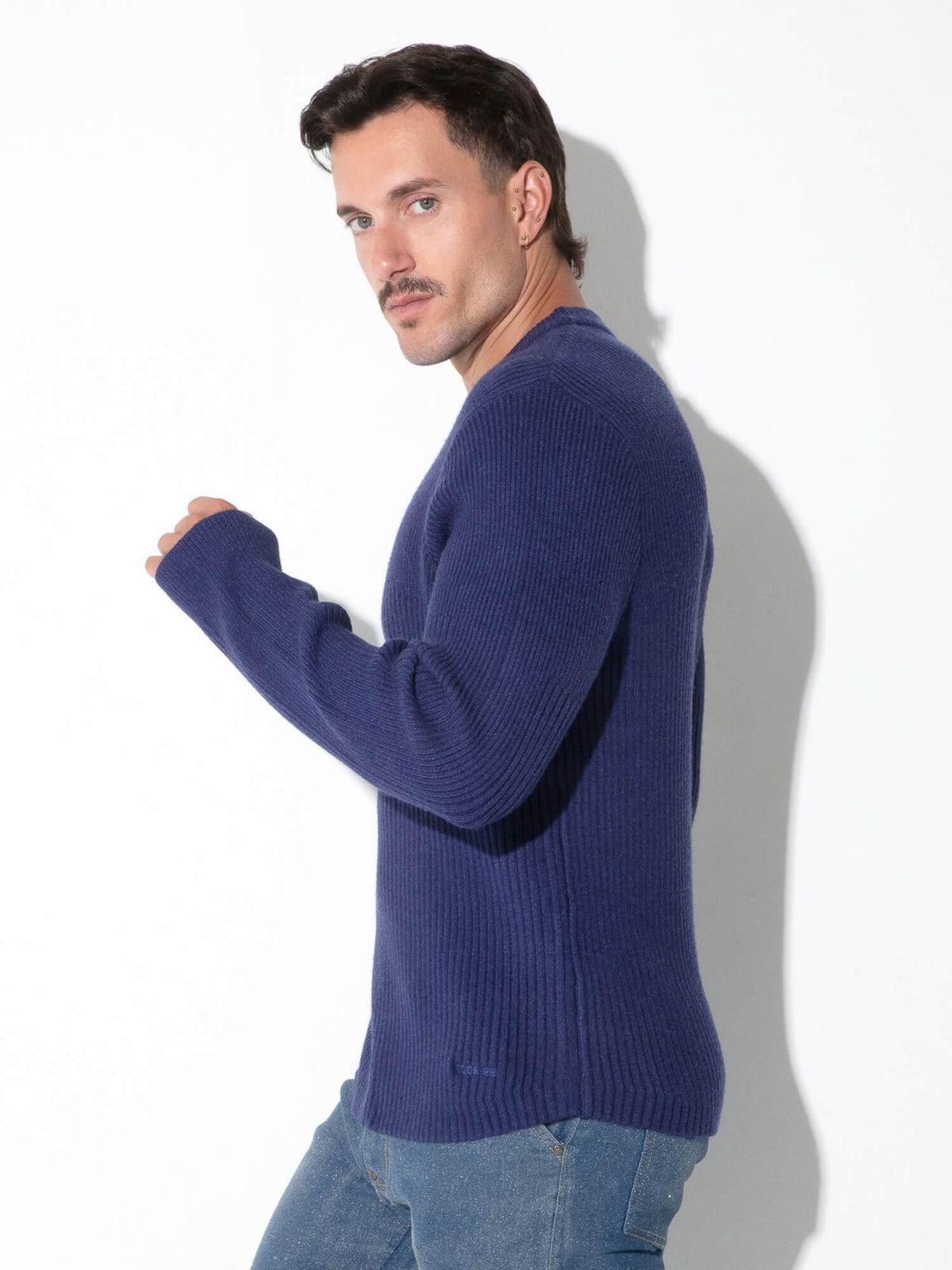 A man wearing a navy blue ribbed knit sweater and light blue jeans, posing against a white studio background with a soft shadow.