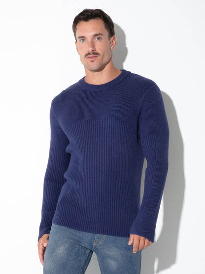 A man wearing a navy blue ribbed knit crewneck sweater and blue jeans, standing against a white studio background with a soft shadow.
