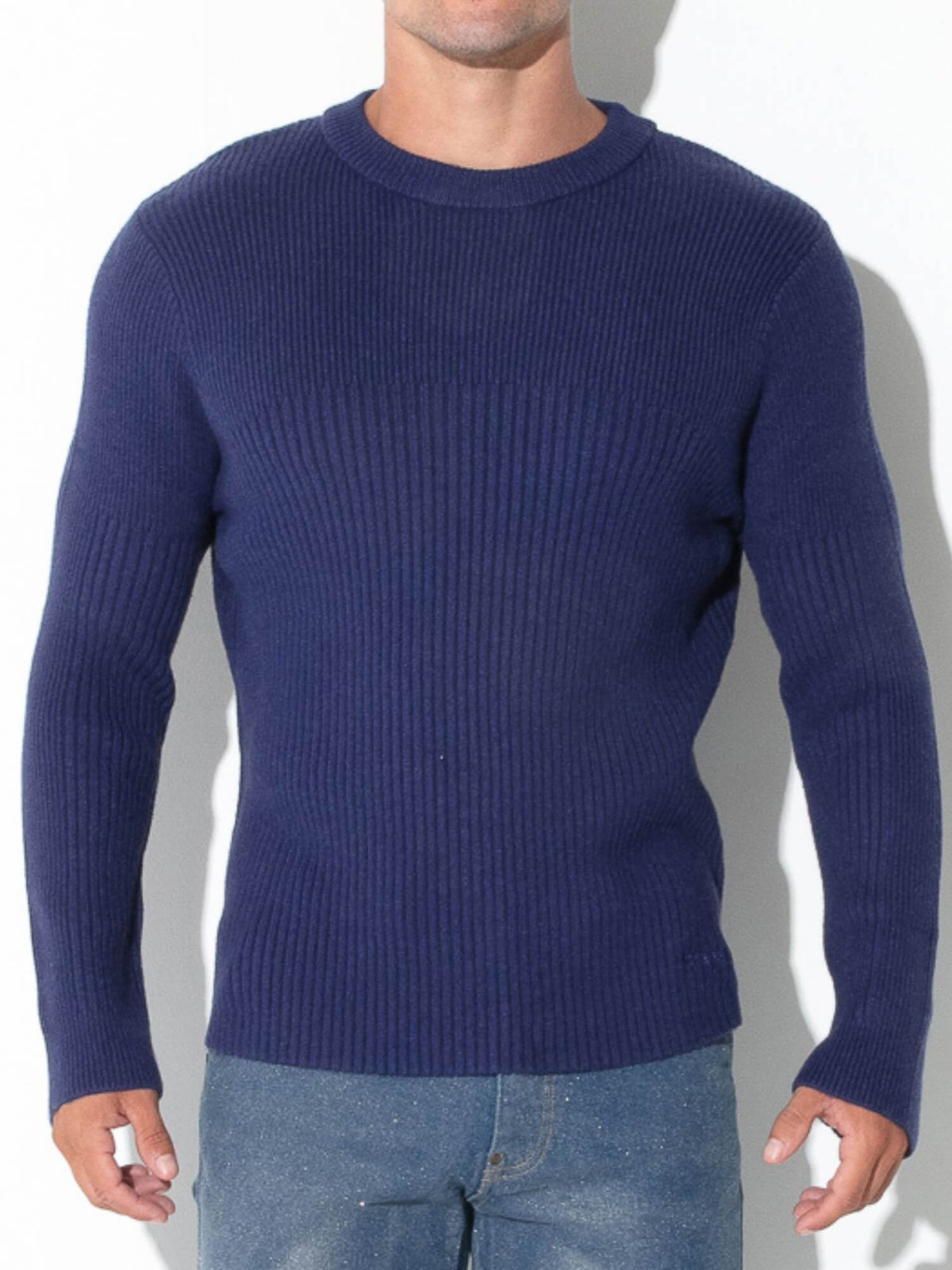 A man wearing a navy blue ribbed knit crewneck sweater and blue jeans, standing against a white background with a soft shadow.