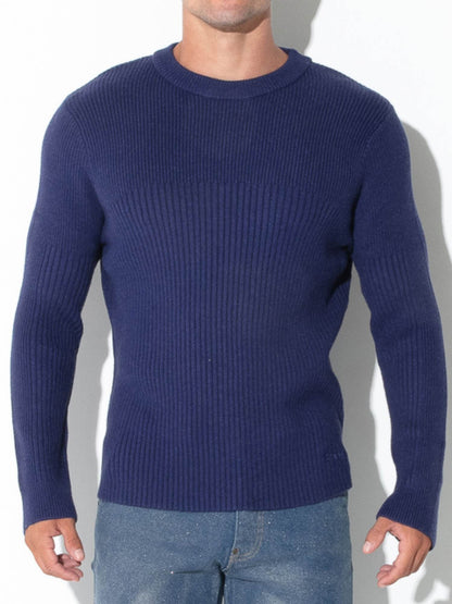 A man wearing a navy blue ribbed knit crewneck sweater and blue jeans, standing against a white background with a soft shadow.