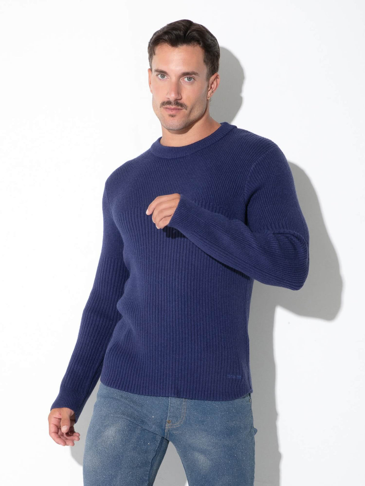 A man wearing a navy blue ribbed knit sweater and blue jeans, standing against a white wall with a soft shadow.