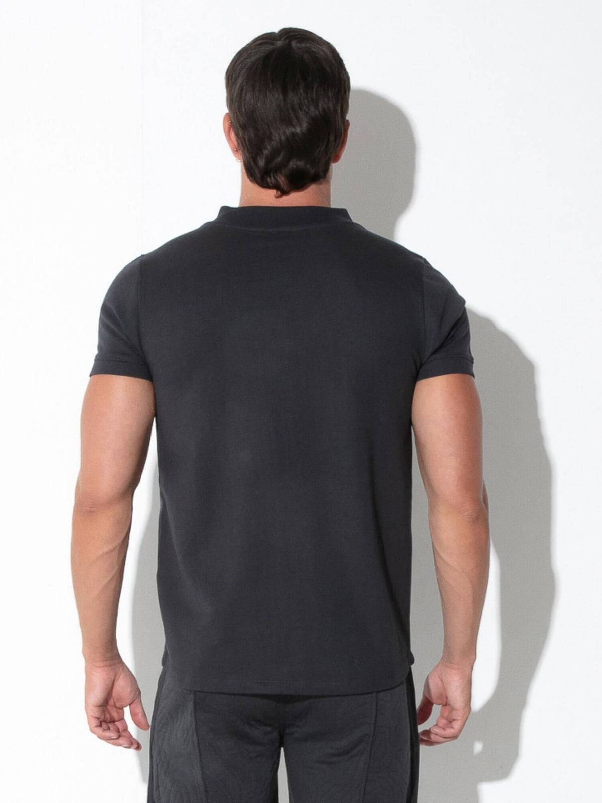 A man viewed from behind wearing a dark charcoal gray short-sleeve polo shirt and matching dark trousers, standing against a white wall with a soft shadow.