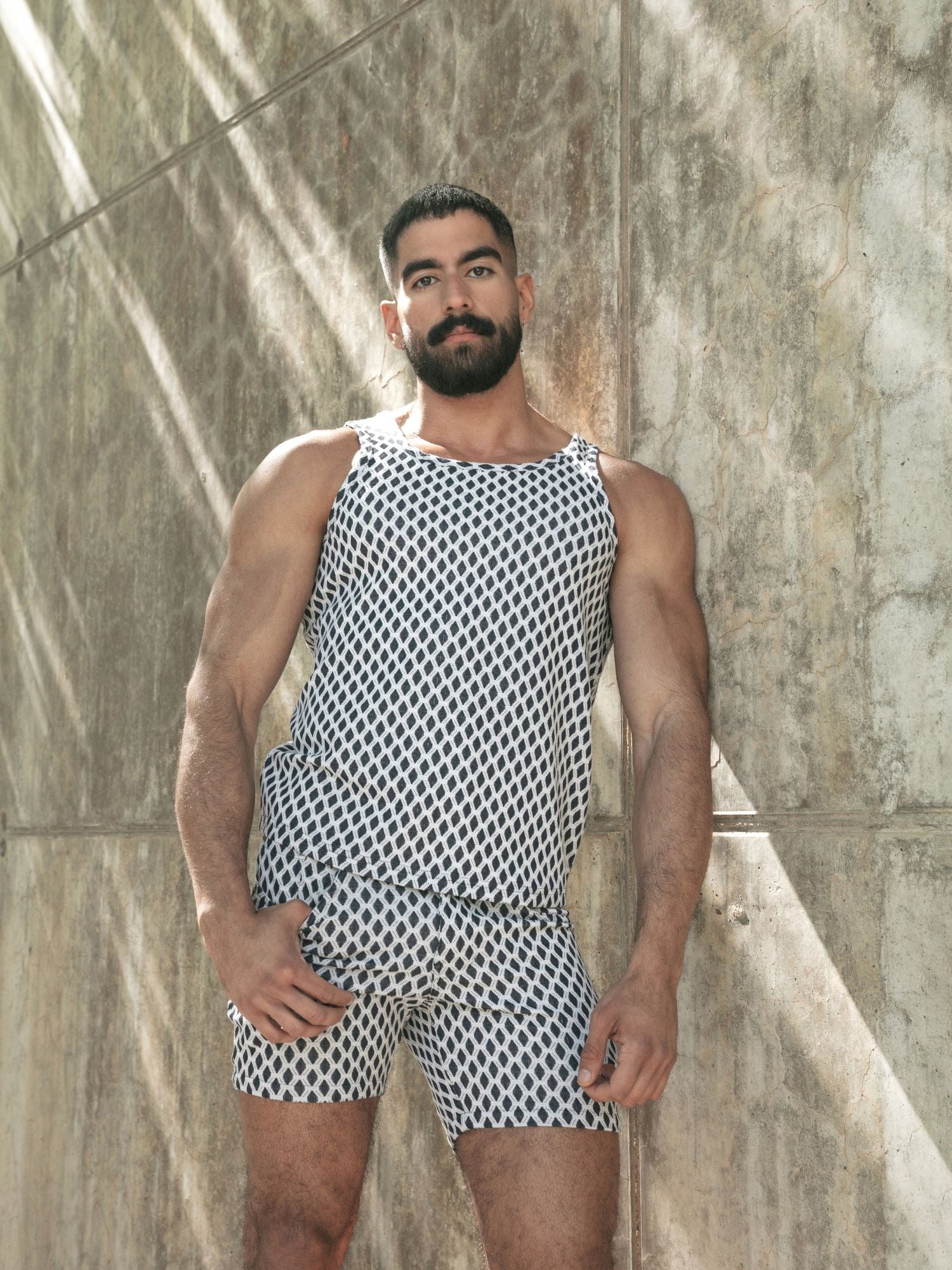 A muscular man with a beard stands against a textured concrete wall wearing a sleeveless tank top and matching shorts set in a black and white geometric diamond pattern.