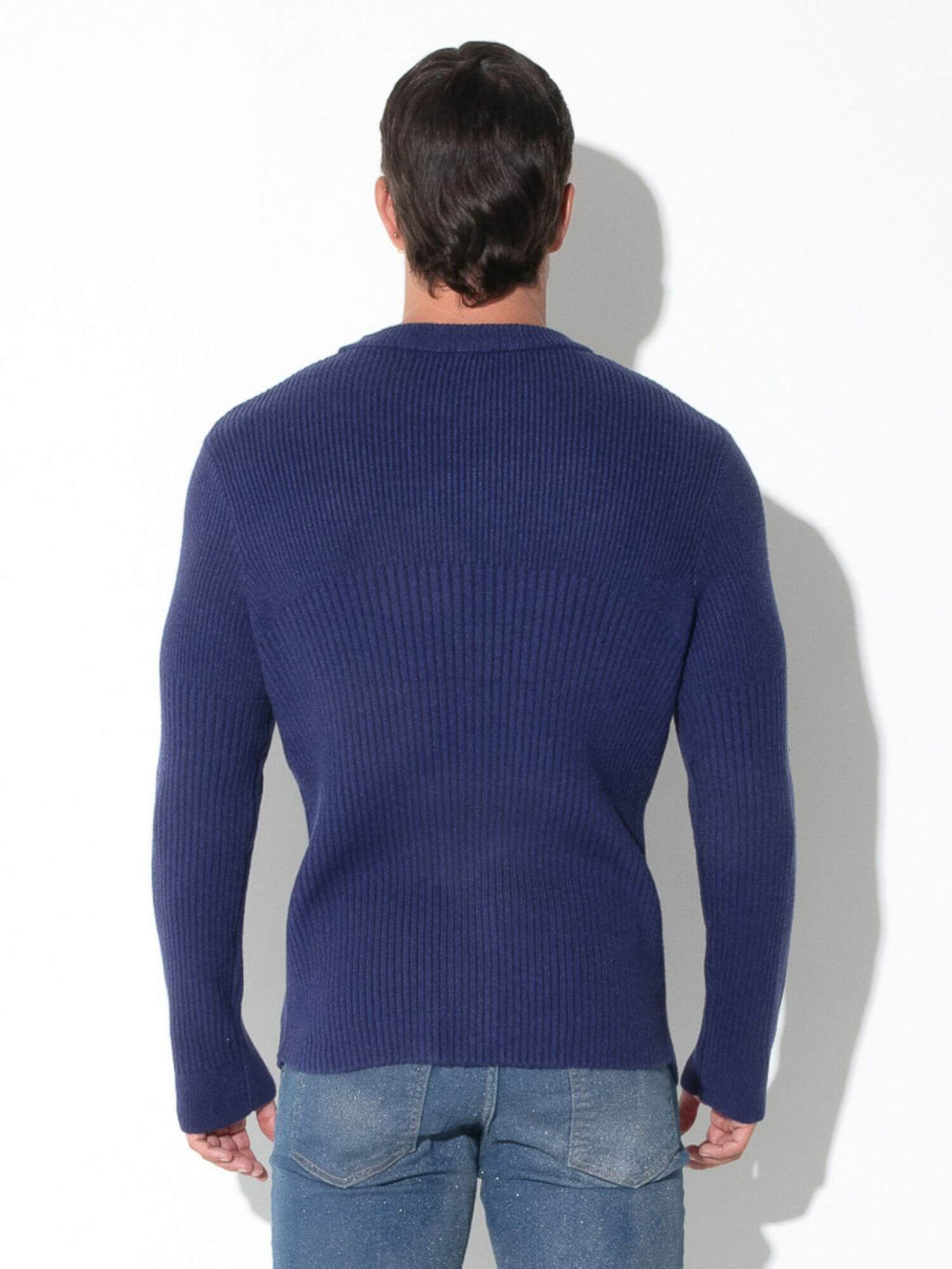 A man viewed from behind wearing a navy blue ribbed knit sweater and blue jeans, standing against a white wall.