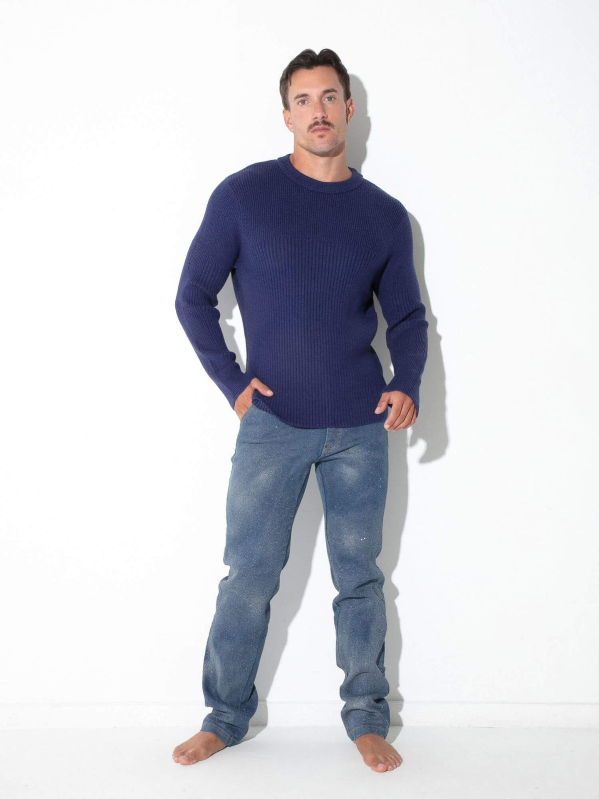 A man wearing a dark navy blue ribbed knit sweater and blue jeans, standing barefoot against a plain white studio background.