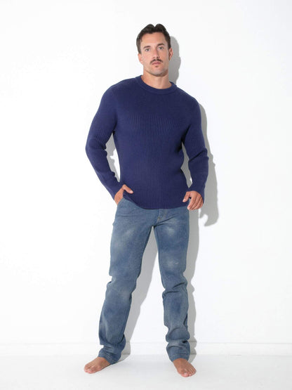 A man wearing a dark navy blue ribbed knit sweater and blue jeans, standing barefoot against a plain white studio background.