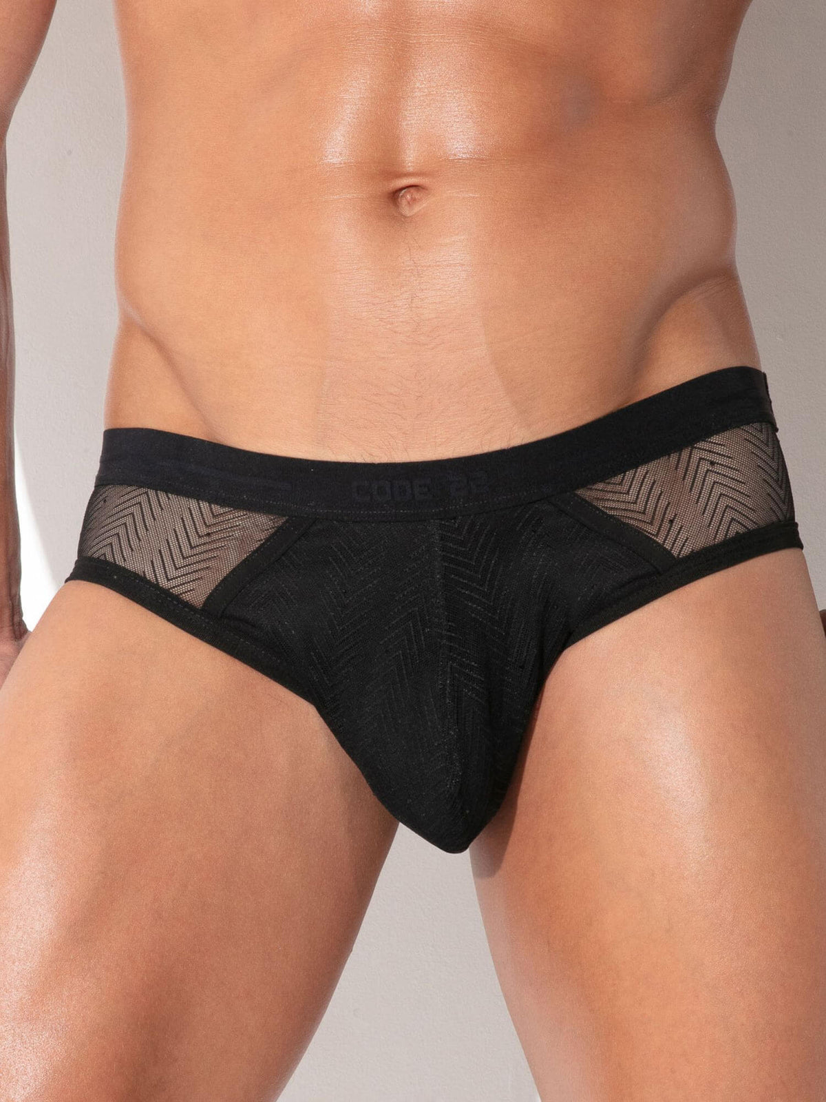 A close-up of a male model's lower torso wearing black men's briefs with sheer chevron-patterned mesh side panels and a black waistband.