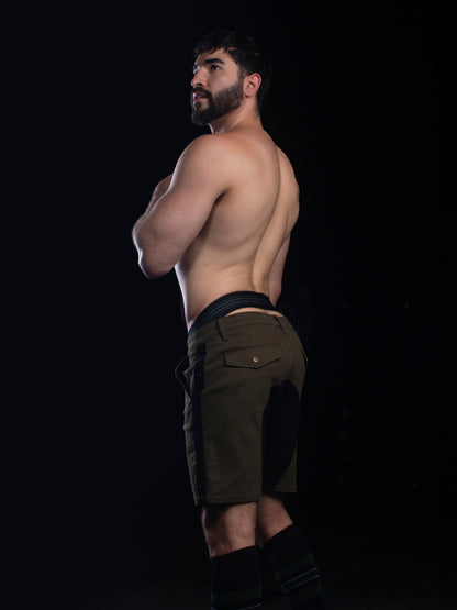 A shirtless man with a beard stands in profile against a black background, wearing olive green athletic shorts with black side panels and black knee-high socks.