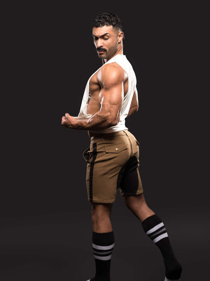 A muscular man posing in a white tank top, brown cargo shorts with black side panels, and black athletic socks with white stripes, set against a solid black studio background.