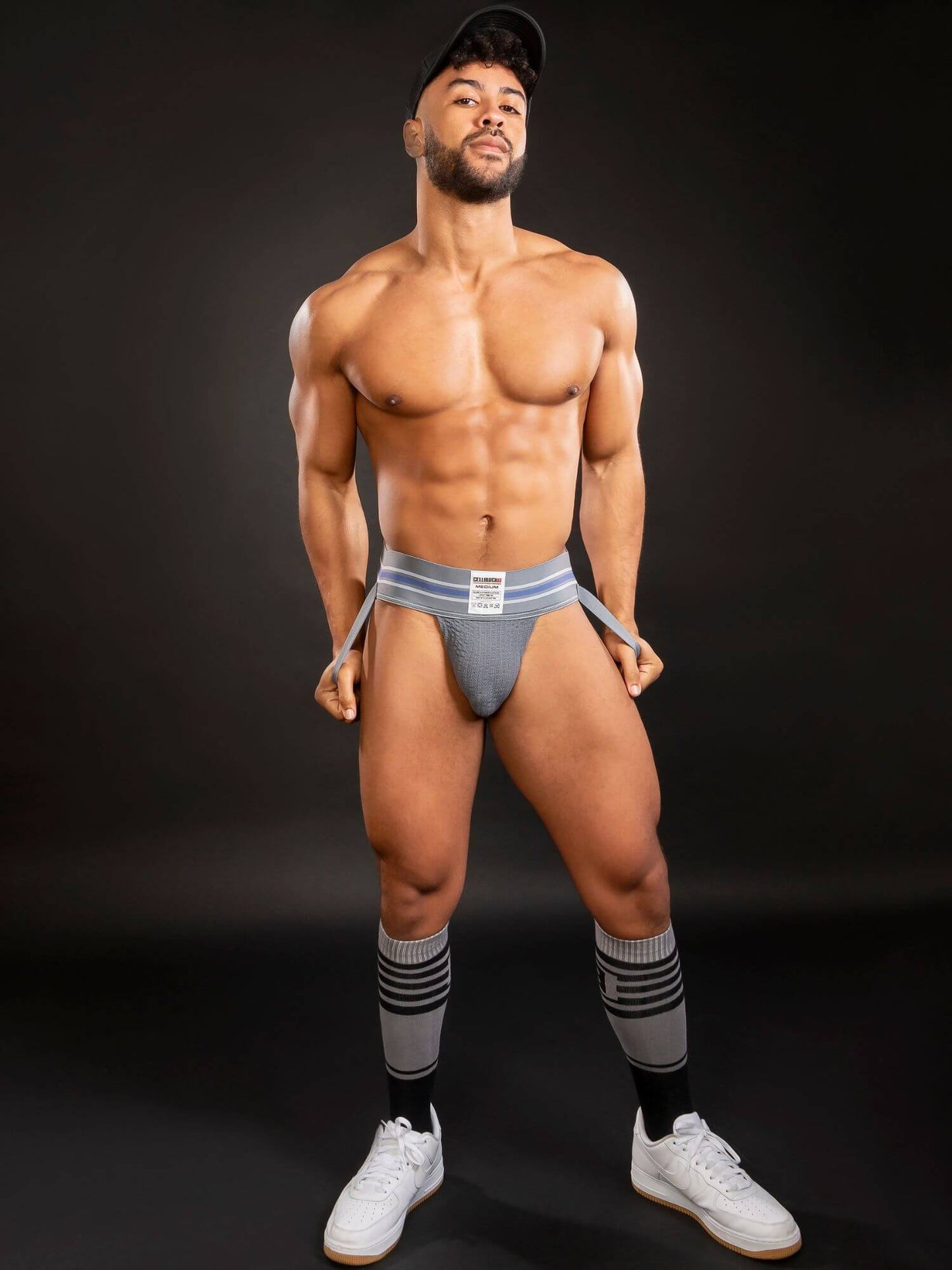 Man wearing gray jockstrap underwear, grey socks and white sneakers on a black background