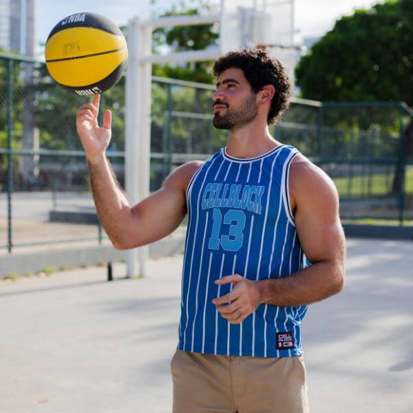 A man on an outdoor basketball court wearing a blue and white pinstriped basketball jersey with 'CELLBLOCK 13' on the front, balancing a yellow and black basketball on his finger.