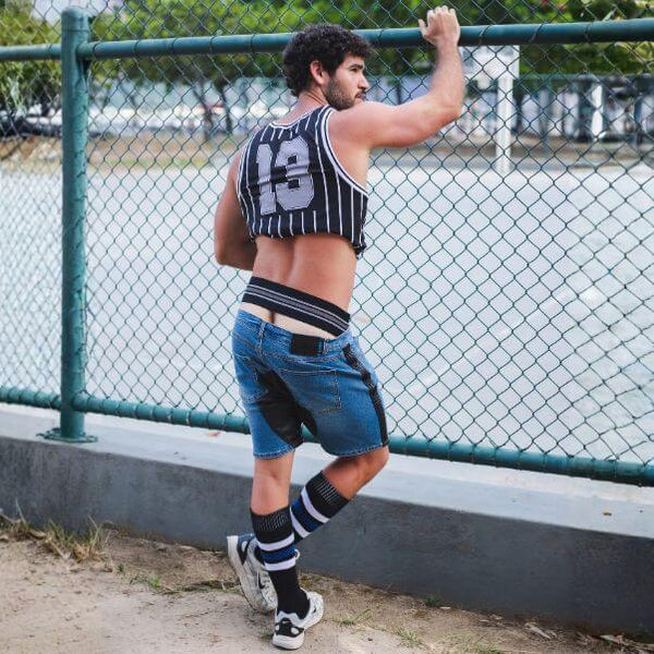 A man wearing a black and white striped tank top with the number 13 on the back, blue denim shorts with black patches, and striped knee socks, posing outdoors in front of a green chain-link fence.