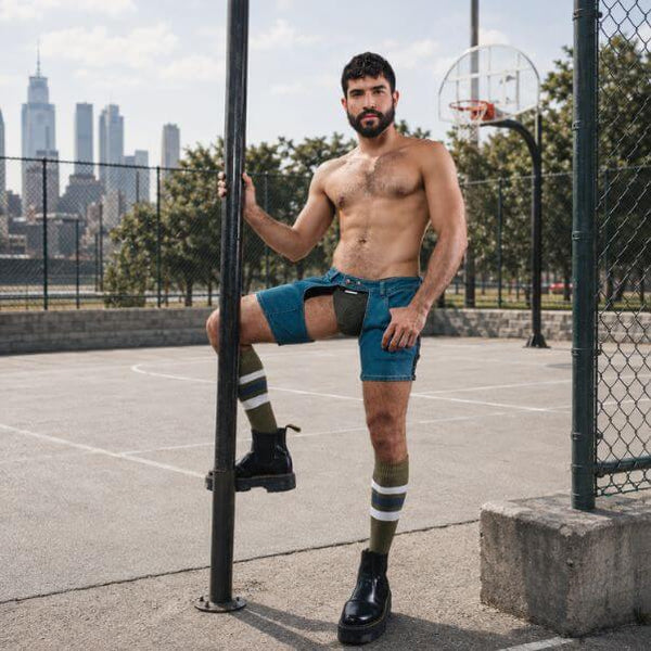 A shirtless man with a beard stands on an outdoor basketball court, wearing teal denim chaps shorts with a jockstrap, striped socks, and black boots, leaning against a black pole.