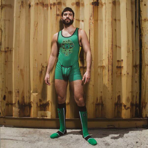 A man wearing a green wrestling singlet with a geometric chest graphic and matching green knee-high socks, standing in front of a rusty corrugated metal shipping container.