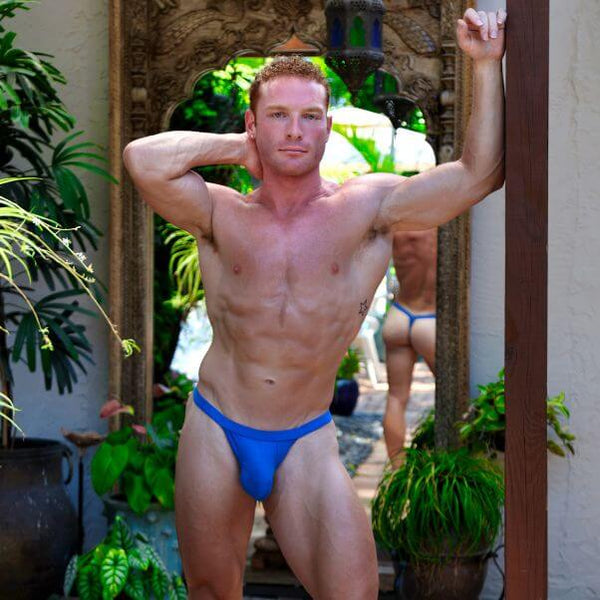 A muscular man posing in a doorway wearing a bright blue SUKREW Fiesta bubble thong.