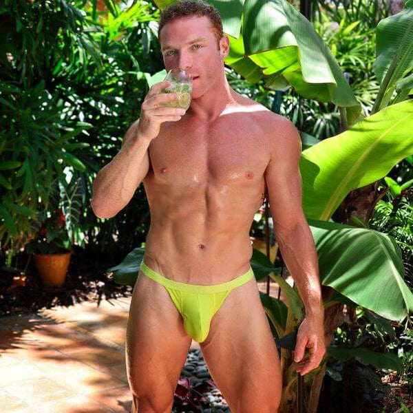 A muscular man standing outdoors in a tropical garden setting, wearing a SUKREW Lime thong and holding a mojito cocktail.