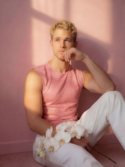 Man in pink tank top and white pants sitting against a pink wall with white flowers.