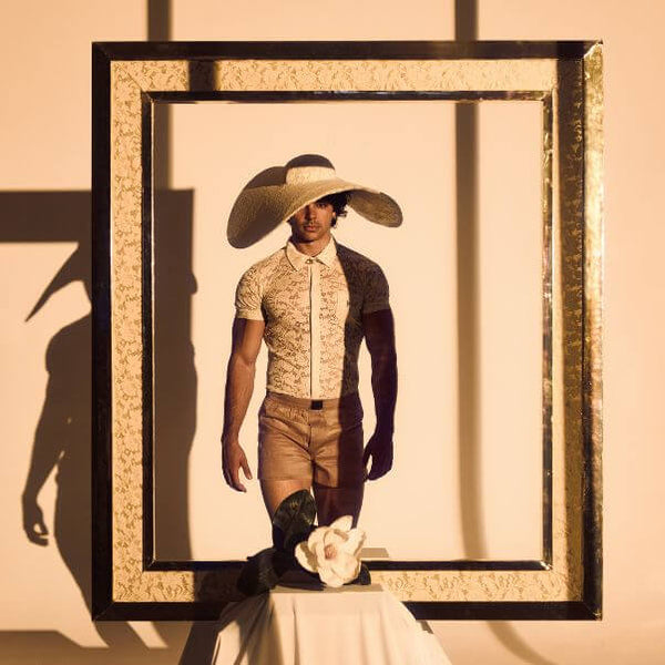 Person wearing a wide-brimmed hat and patterned shirt framed by a decorative black and gold frame on a beige wall.