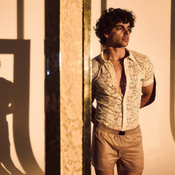 Man wearing a patterned shirt and beige shorts standing against a textured wall with shadows.
