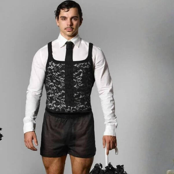 Man wearing a white shirt, black lace suspenders, and brown shorts against a gray background