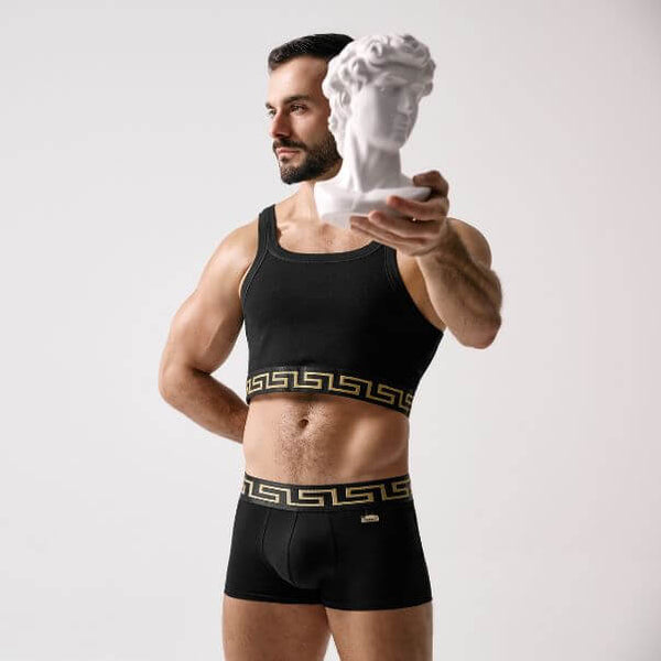 Man wearing black underwear with a decorative waistband, holding a white bust against a plain background.