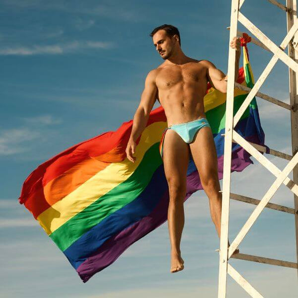 Man holding a rainbow flag against a blue sky wearing MODUS VIVENDI Pride swimwear