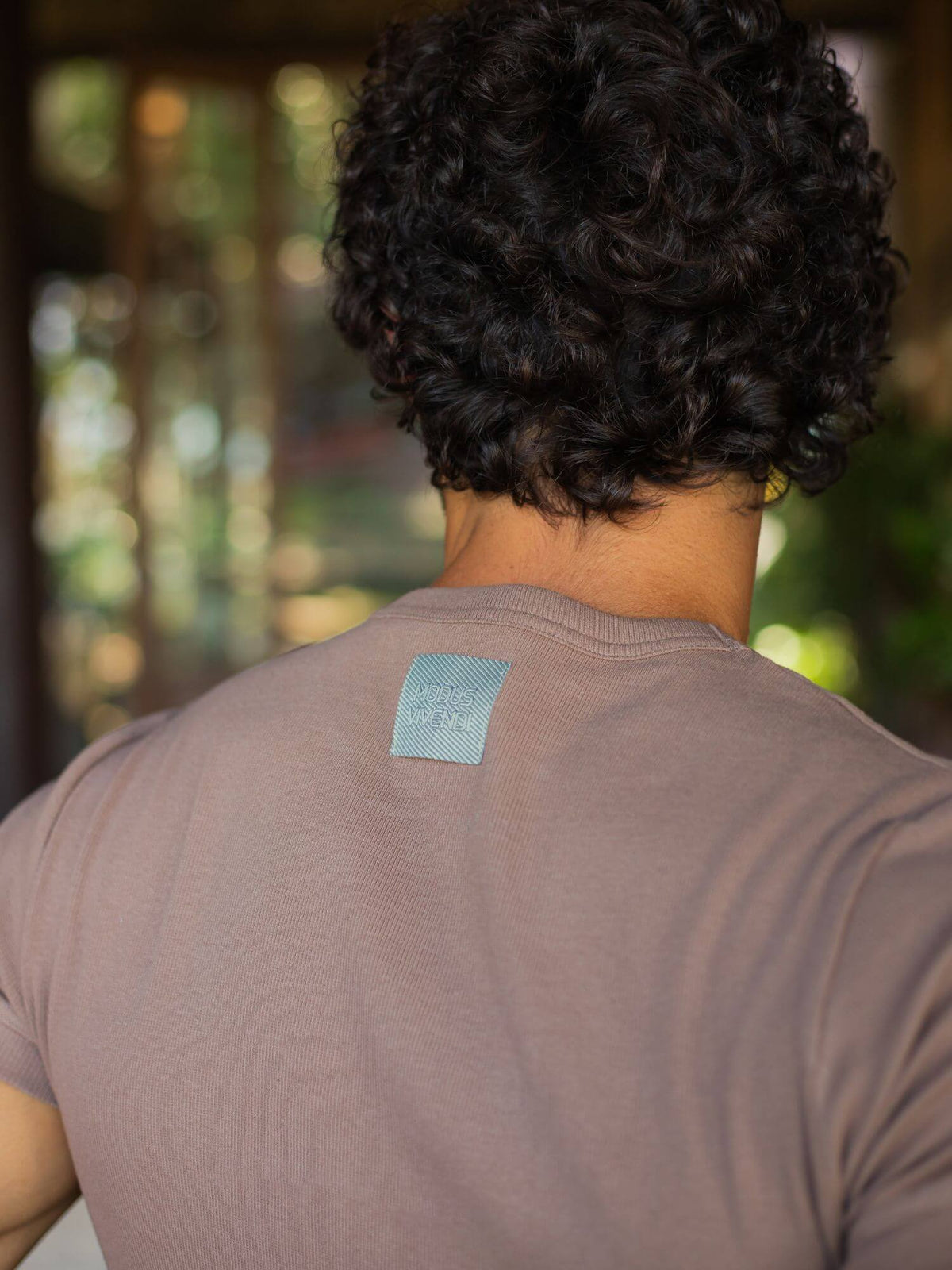 Back view of a person with curly dark hair wearing a taupe-brown short-sleeve t-shirt with a small light blue square patch on the upper back, set against a blurred outdoor background.