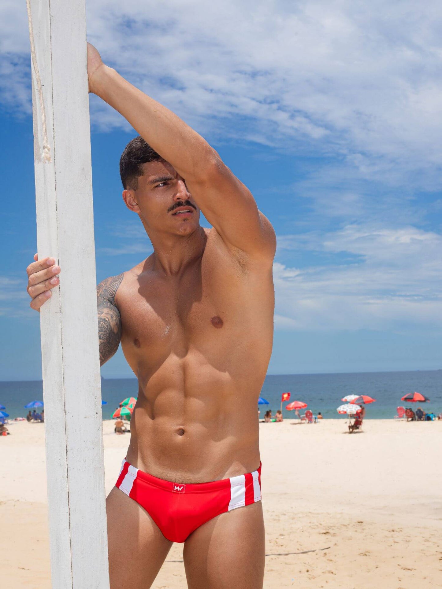 Man in red swim brief holding a pole on a beach with people and umbrellas in the background.