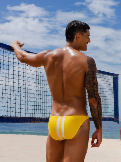 Man in yellow swim brief standing on a beach with a volleyball net in the background.