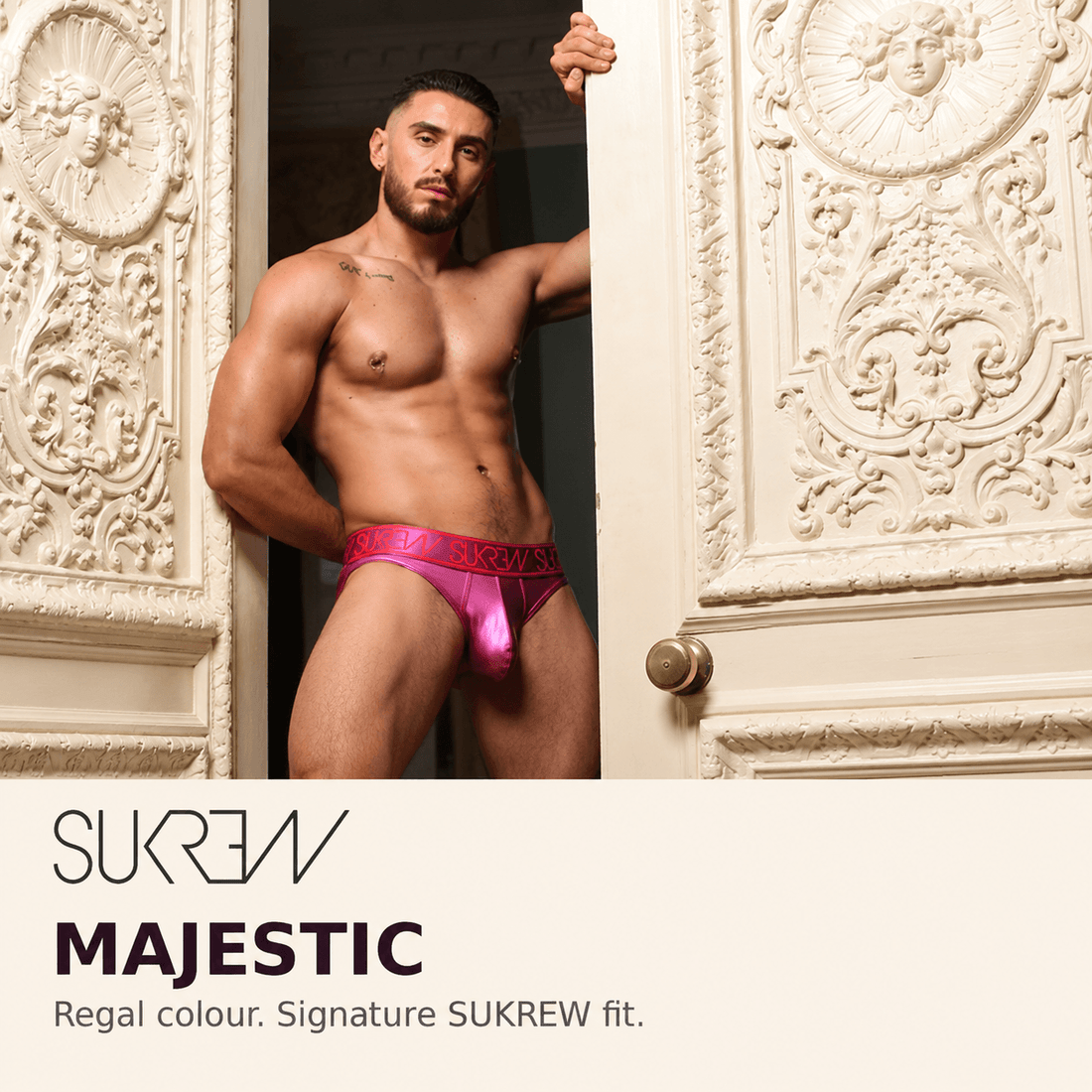 A shirtless male model wearing bright pink sukrew satin briefs with a red patterned waistband, posed between two ornate carved cream doors. Text at the bottom reads 'SUKWAV MAJESTIC Regal colour.