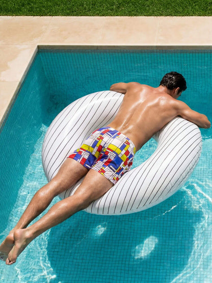 A man floating face-down in a swimming pool on a large white striped inflatable ring, wearing colorful geometric print Modus Vivendi Mondrian swimming shorts.