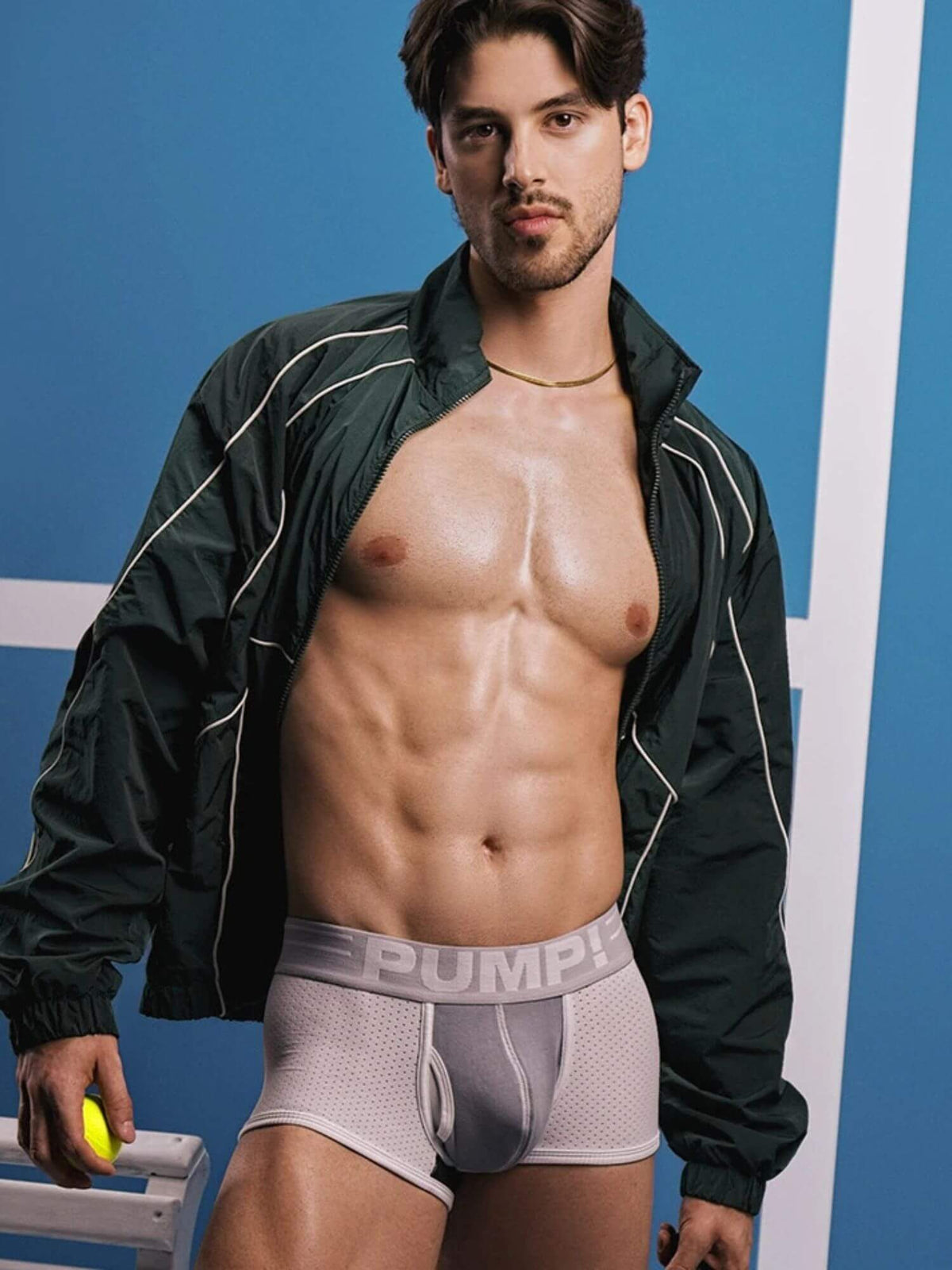 A male model wearing PUMP! brand light gray mesh boxer briefs, with his dark green zip-up jacket open, posing against a blue and white background.