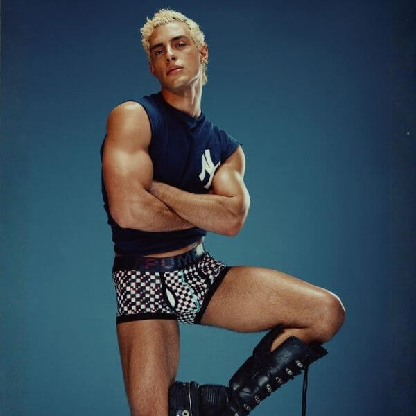 Man posing in PUMP! Glitch checkered boxer trunks and black boots against a blue background