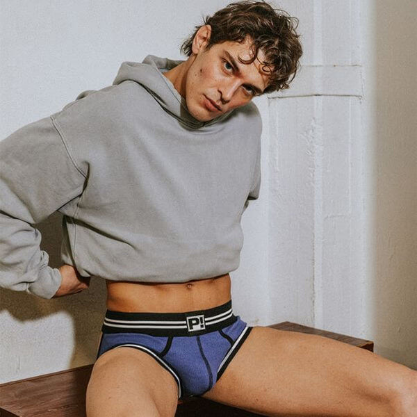 Person wearing a gray hoodie and blue PUMP! Round2 underwear with a logo, sitting against a white wall.