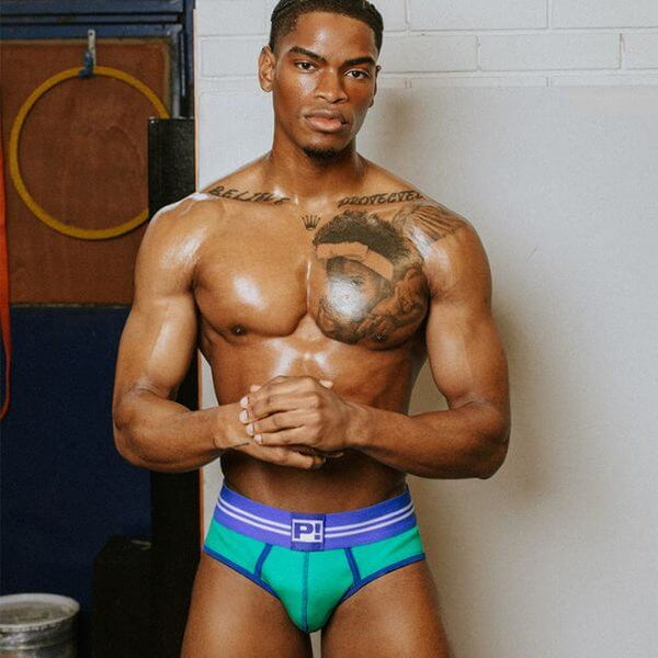 Man wearing green and blue PUMP! Round2 underwear with a logo, standing against a white wall.