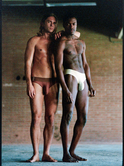 Two men standing barefoot indoors, each wearing minimal underwear. The man on the left wears dark brown briefs, and the man on the right wears white briefs.