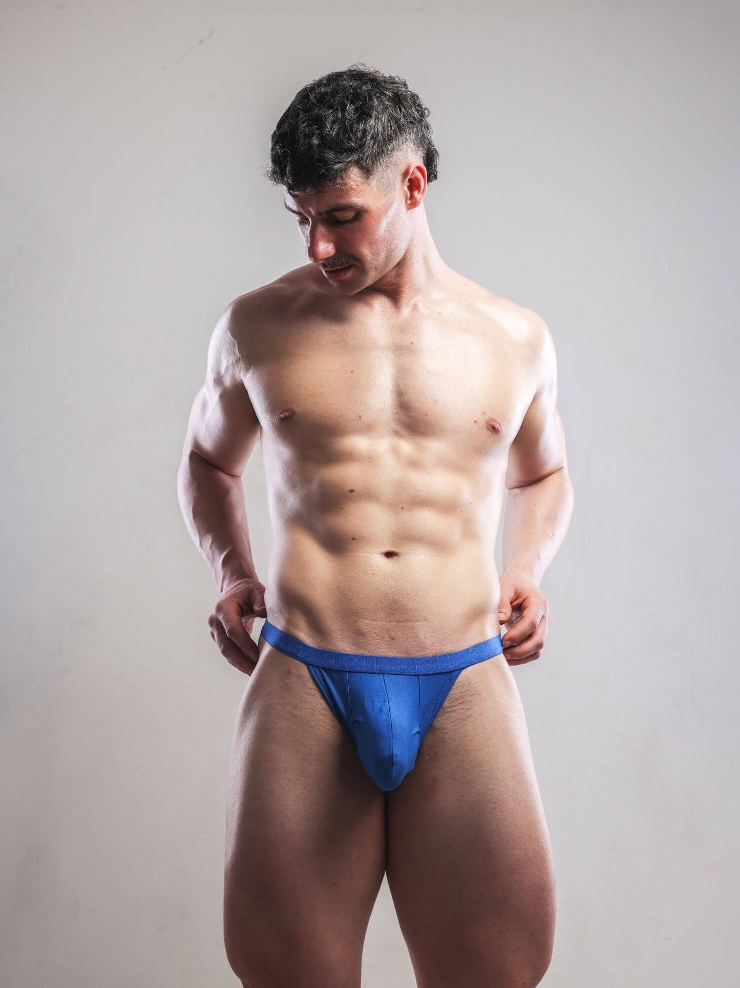 A muscular man wearing a bright blue thong standing against a plain light gray studio background.