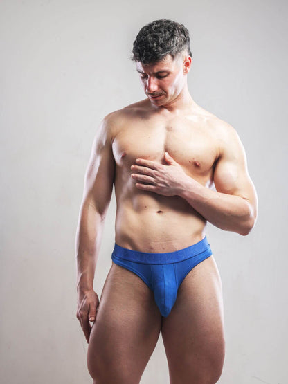 A muscular man wearing a bright blue men's thong underwear bottom, standing against a plain light gray studio background.