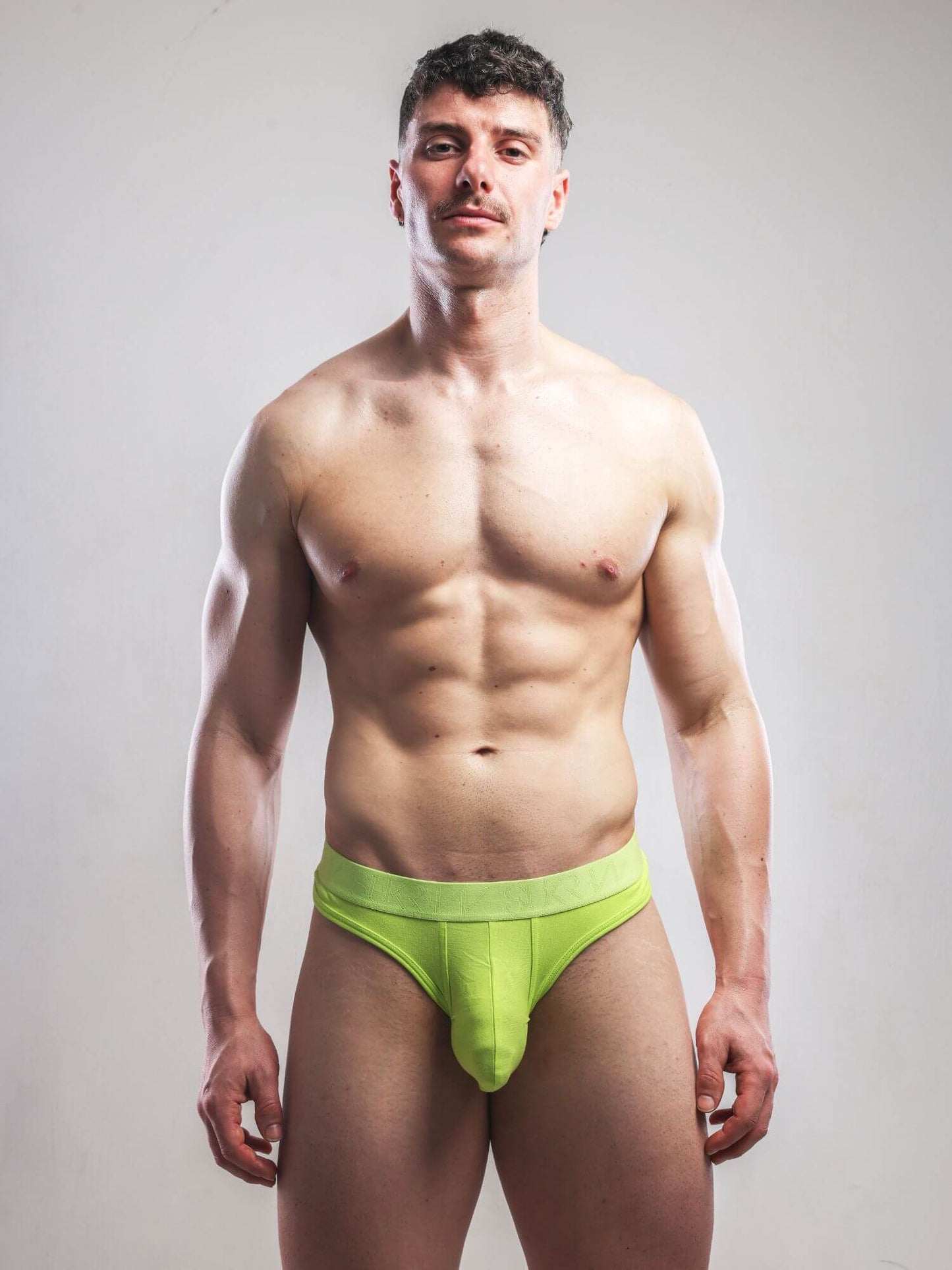 A muscular man wearing a bright lime green men's thong, standing against a neutral light gray studio background.