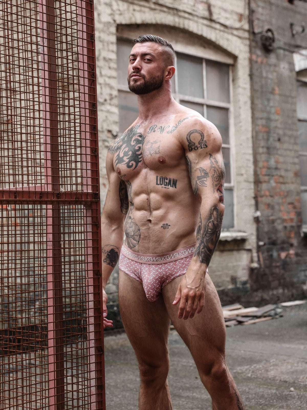 A muscular, tattooed man wearing red patterned briefs. The underwear has a visible waistband with the brand name printed on it.