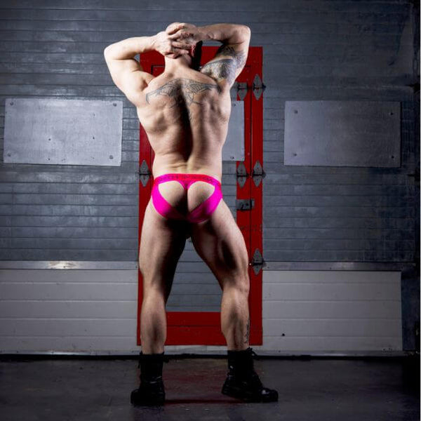 A muscular man posing from behind wearing SUKREW bright hot pink thong-style underwear, standing in front of a gray brick wall with a red door frame.