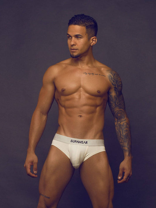 A muscular man with tattoos models white SUPAWEAR briefs against a dark gray studio background.