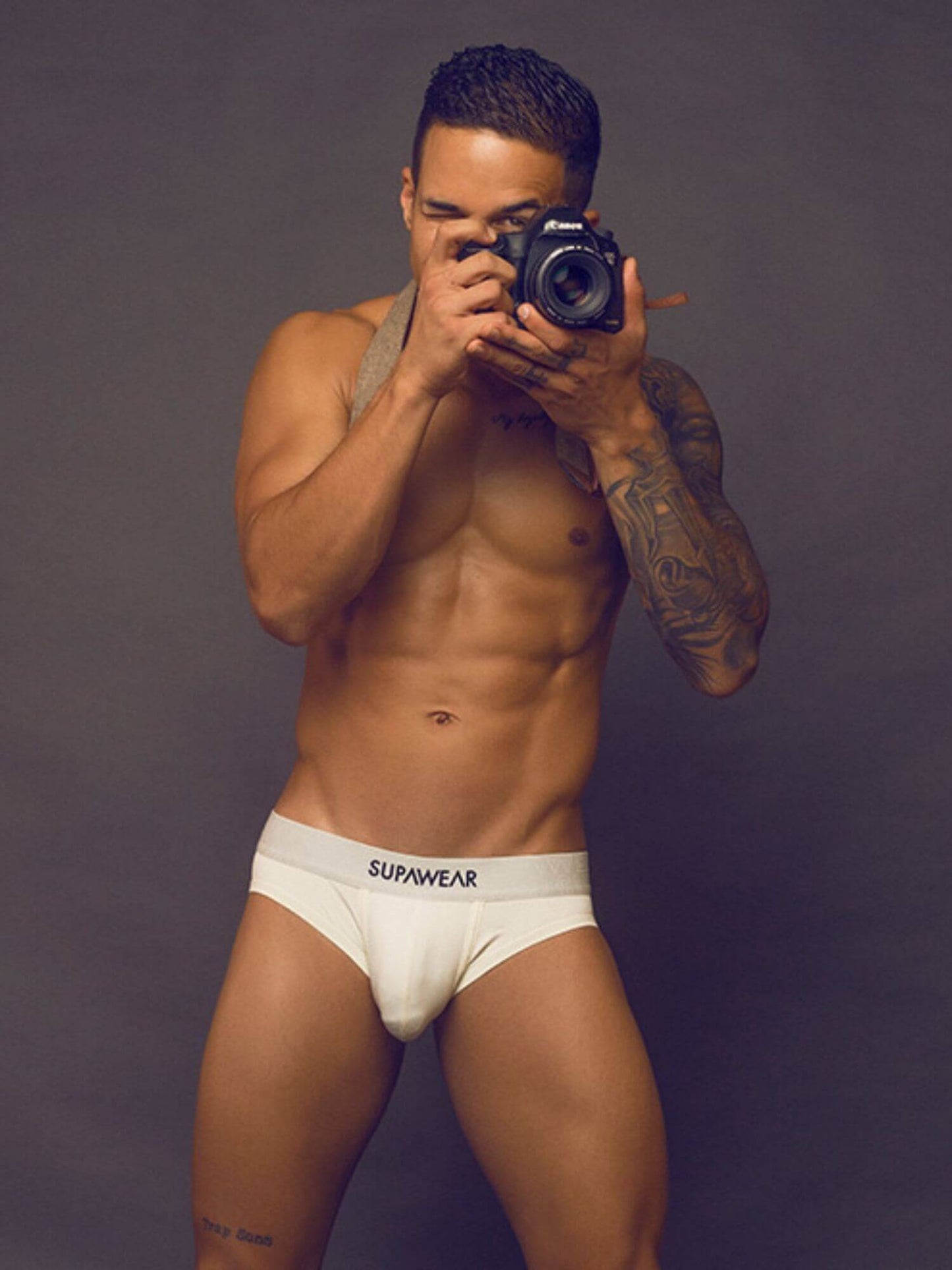 A muscular man posing in a mirror selfie wearing white Supwear briefs, holding a black camera against a dark gray studio background.