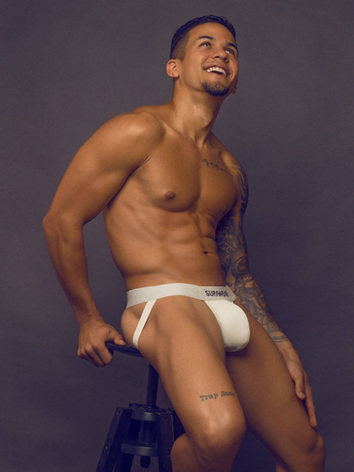 A shirtless man with tattoos wearing white underwear sits on a stool against a dark gray studio background.