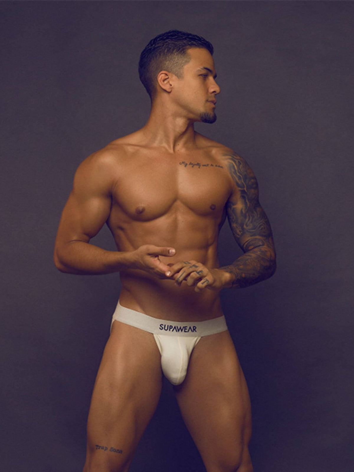 A muscular man wearing white SUPWEAR jockstrap underwear, standing against a dark gray studio background.